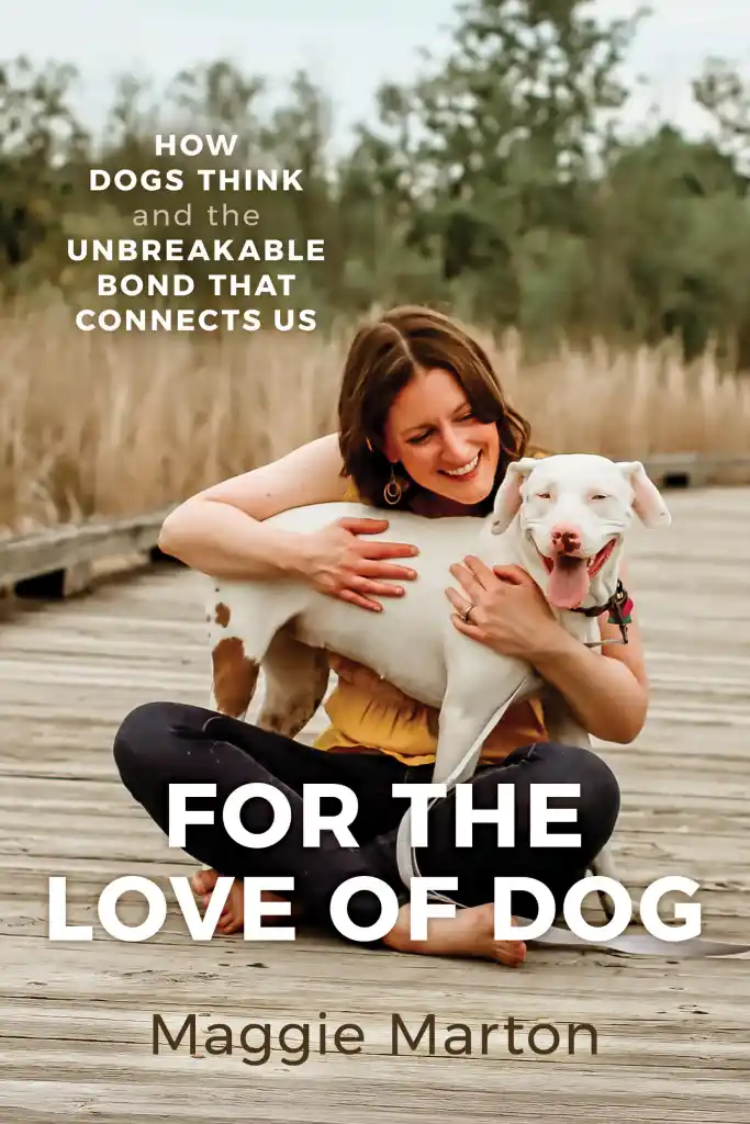 The cover of the book For the Love of Dog shows a white woman with brown hair wearing dark jeans and a yellow blouse sitting on a boardwalk hugging a white dog whose mouth is hanging open with her tongue out.