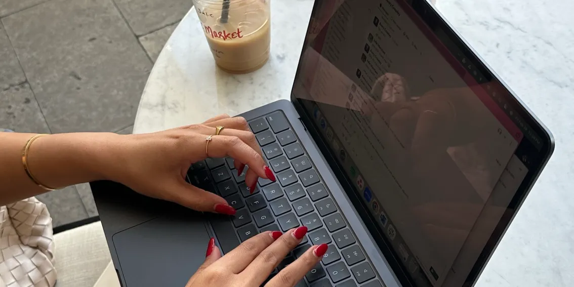 Hands typing on a laptop at an outdoor café table with an iced coffee and water cup nearby, representing the flexibility of writing for blogs and Substack from anywhere.