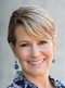 A professional headshot of a woman with short blonde hair, smiling, wearing earrings against a blurred neutral background.